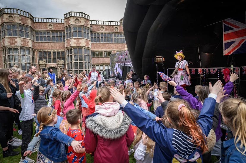 This is an image of Louby Lou the female clown atop a large stage giving an exclusive performance for a crowd of children who are raising their hands in the air.