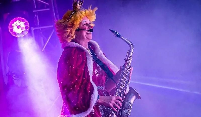 An image depicting Louby Lou the Children’s Entertainer at a show in her clown outfit giving a joyful saxophone performance atop a stage.