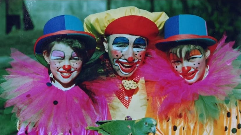 This is an image depicting Louby Lou the Children’s Entertainer during her childhood, another child and her father - all dressed as clowns with heavy colourful makeup and flashy outfits. There is a green parrot in her father's hands.